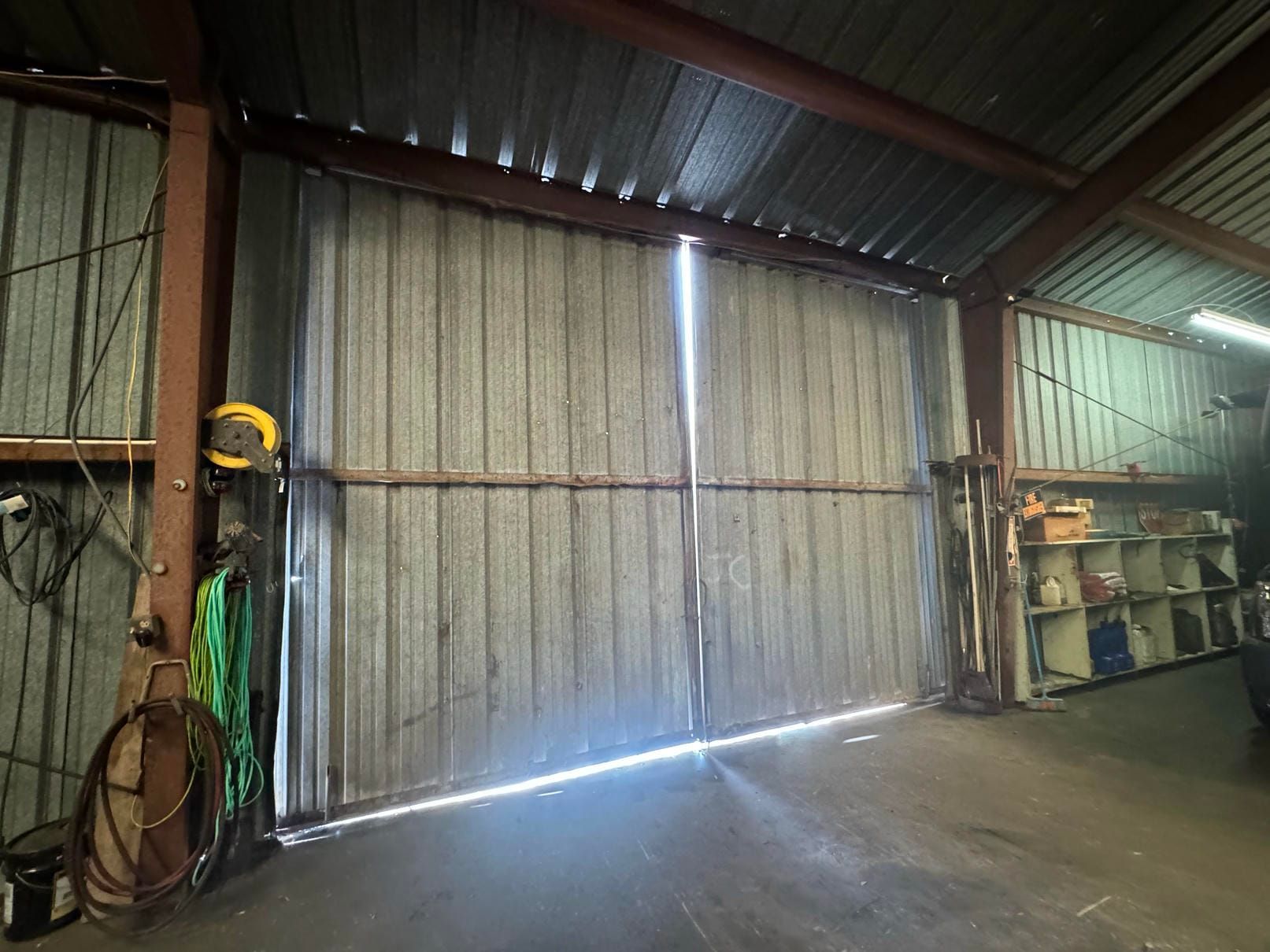 Industrial shed interior with corrugated metal walls, hanging hose, shelves, and bright light under the closed door