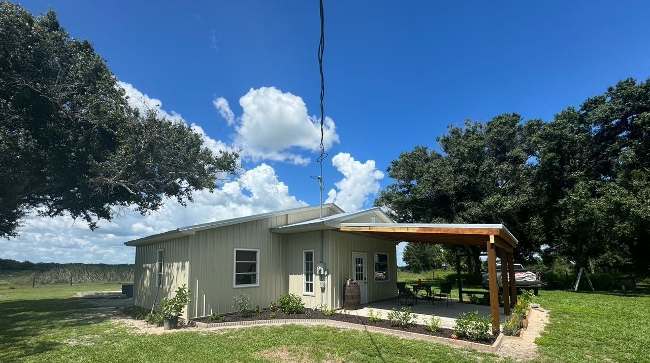 Small house with a carport, set against a backdrop of trees and a bright blue sky with clouds.