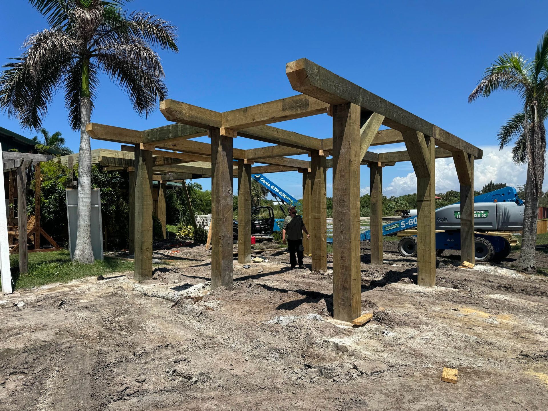 Wooden pergola frame under construction on a sandy site with palm trees and workers nearby