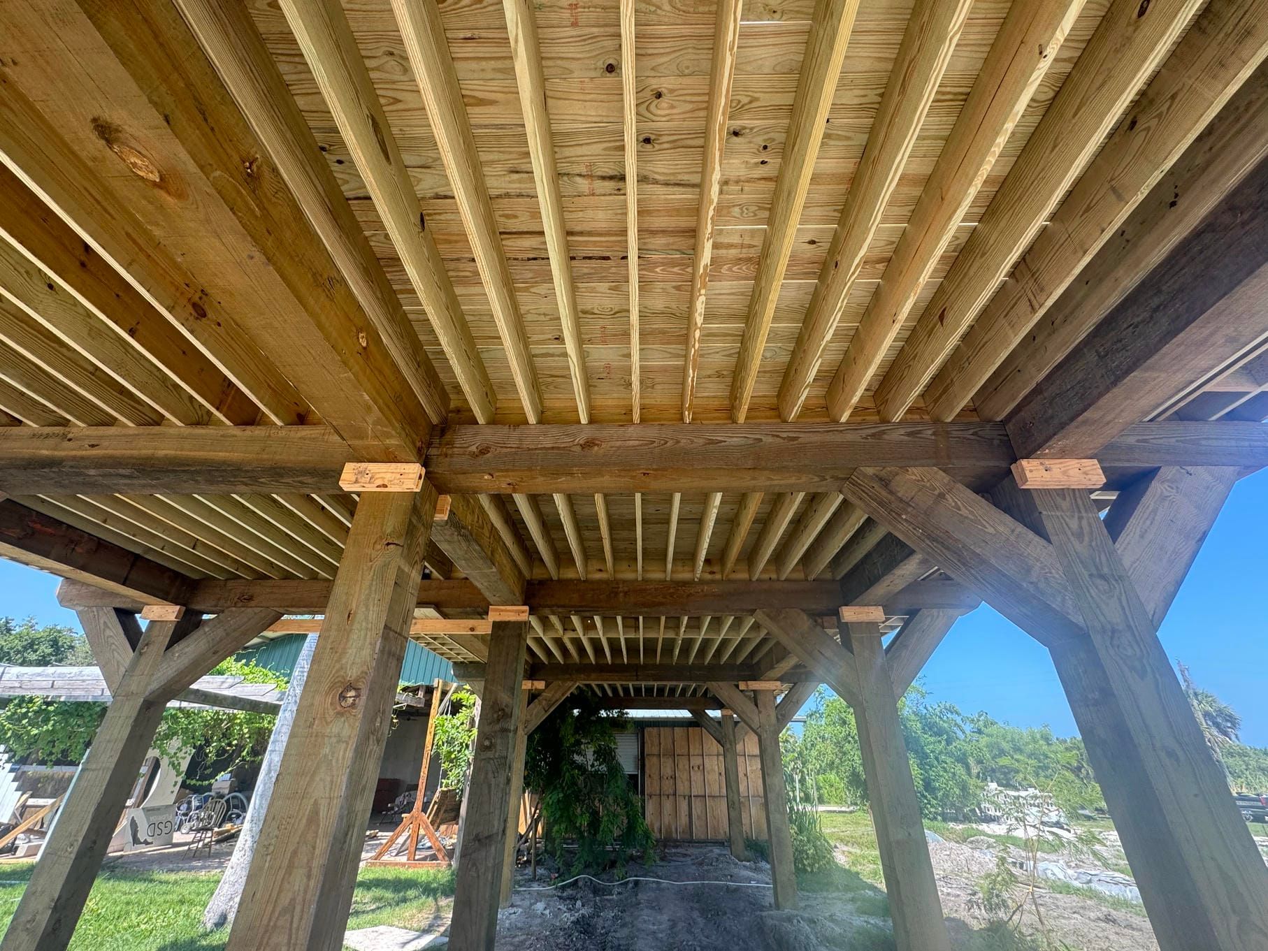 Wooden boardwalk beneath a rustic timber structure, with bright sky and greenery beyond.