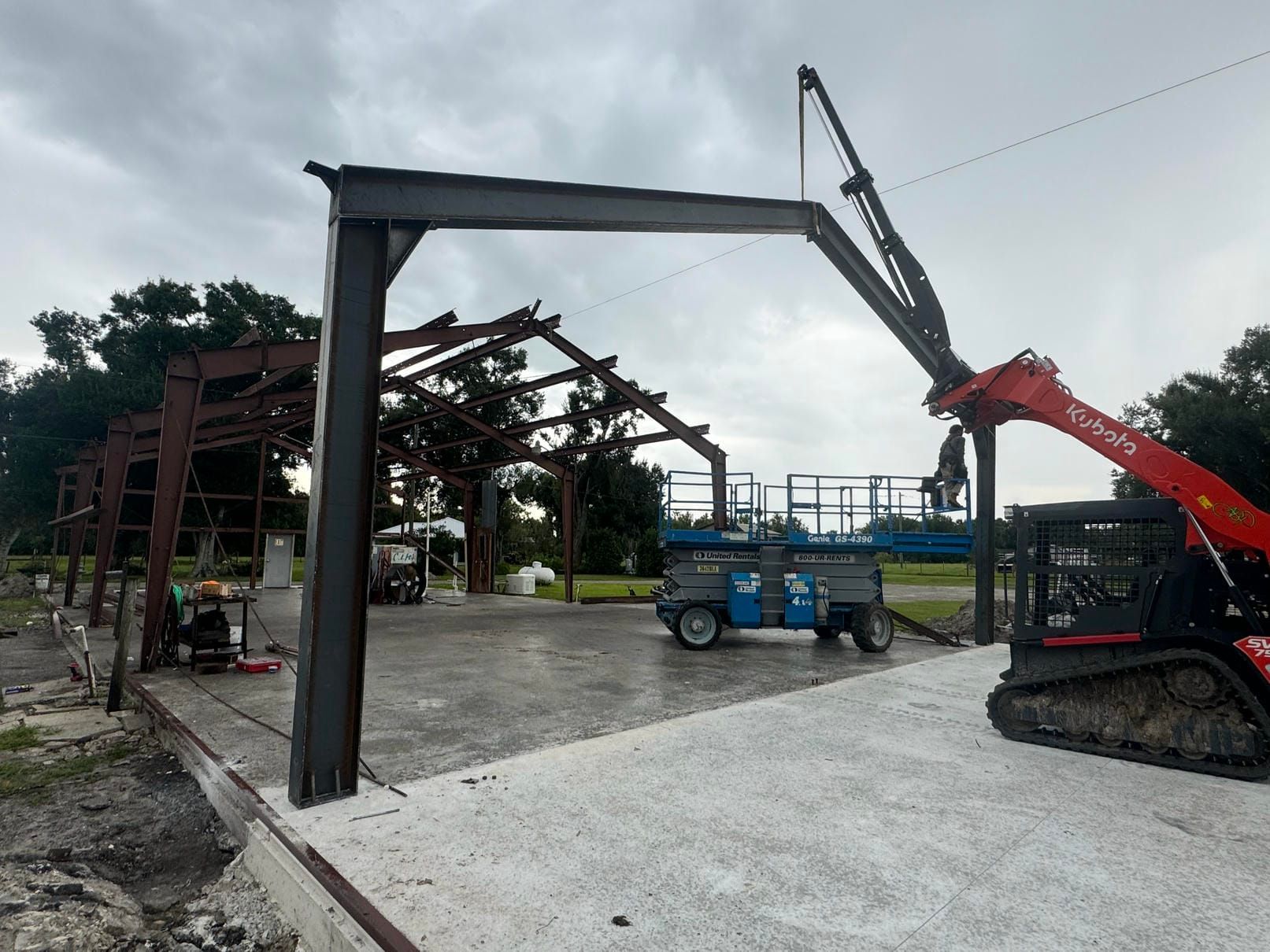 Construction site with steel frame being lifted by an excavator next to a blue lift truck