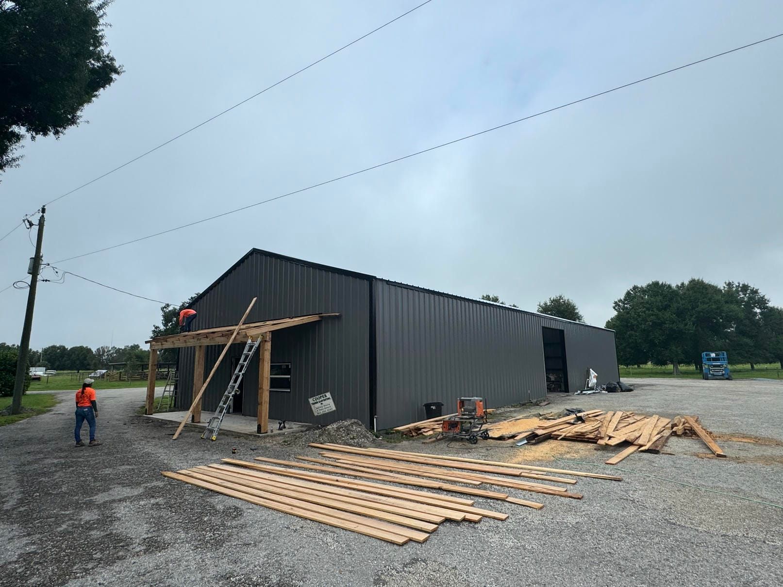 Gray metal building under construction with lumber scattered outside and a worker near the entrance