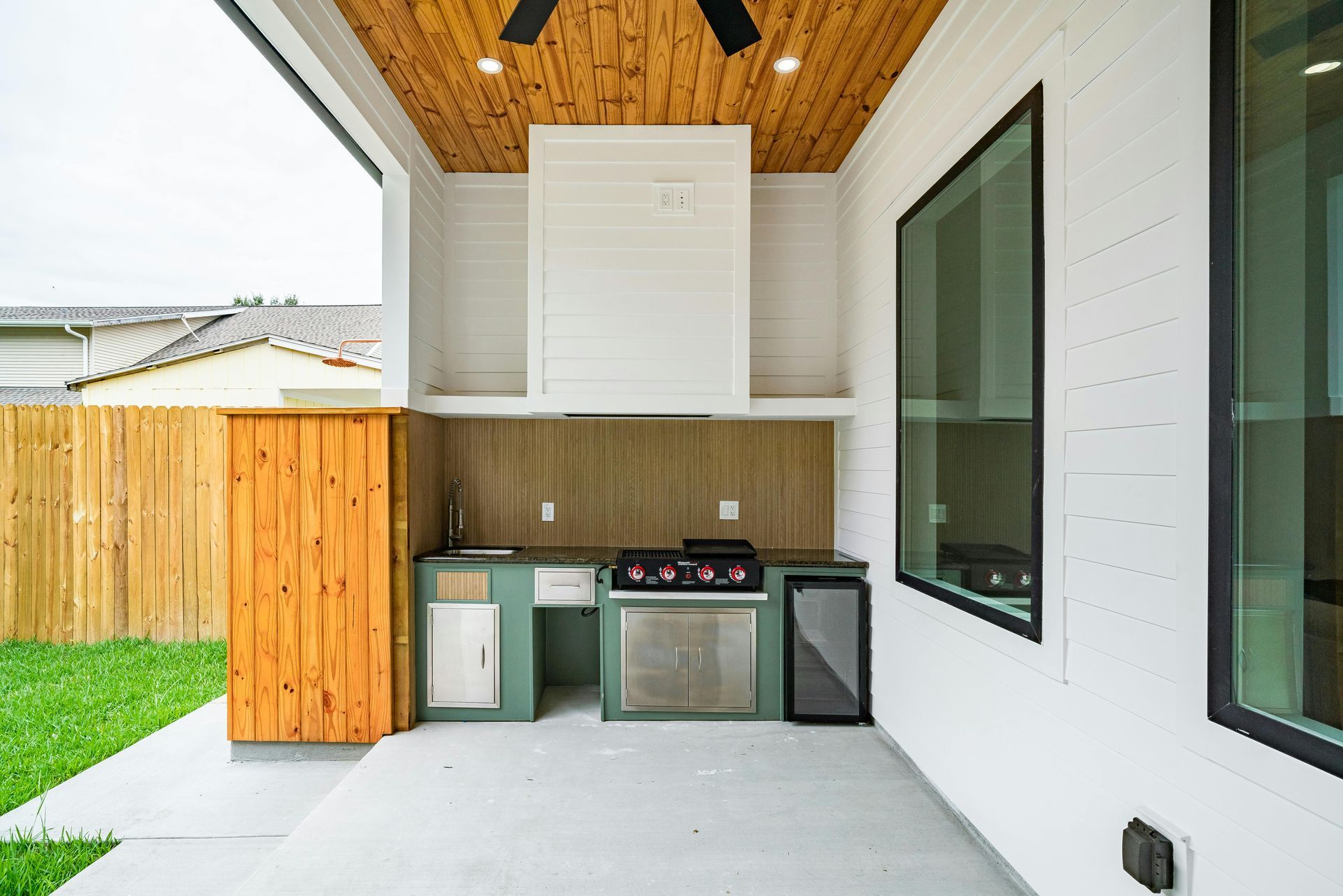 Covered outdoor kitchen with wood ceiling, white cabinets, grill, and sink beside a grassy yard