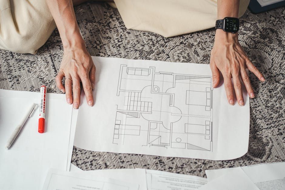 Hands reviewing a floor plan on a carpeted table with a red pen and smartwatch visible.