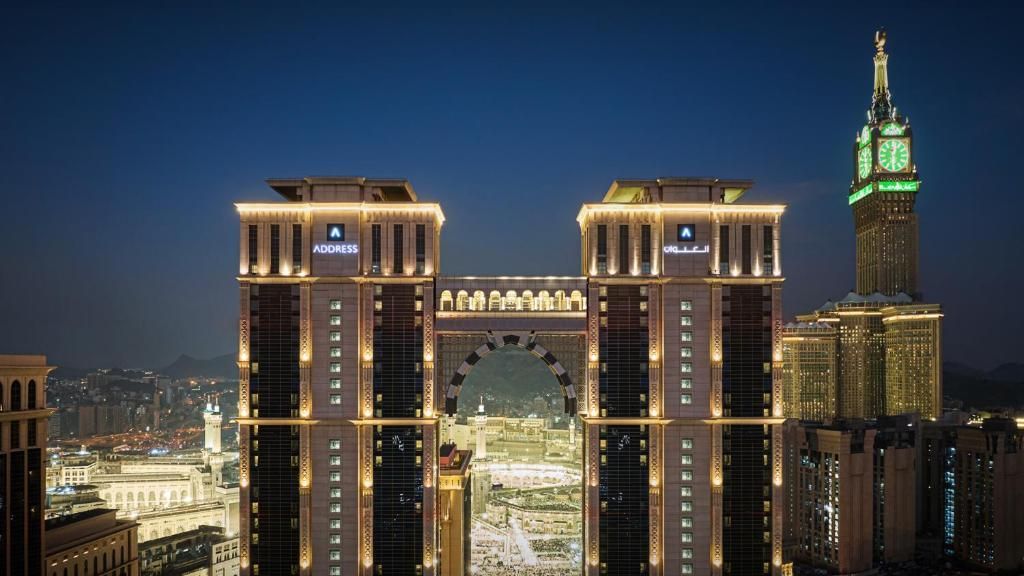 Night view of a modern hotel complex in Mecca, Saudi Arabia, with the illuminated Abraj Al-Bait Clock Tower in the background.