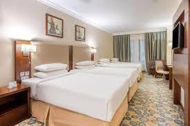 Hotel room with three beds, beige walls, artwork, and patterned carpet.