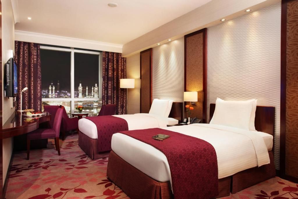 Hotel room with two twin beds, burgundy accents, window view, and desk.