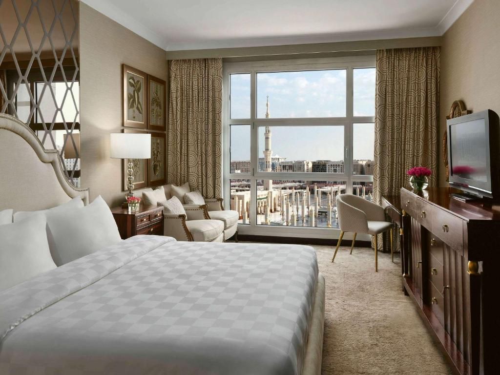 Bedroom with large window overlooking a mosque; bed, seating area, desk, brown and beige tones.