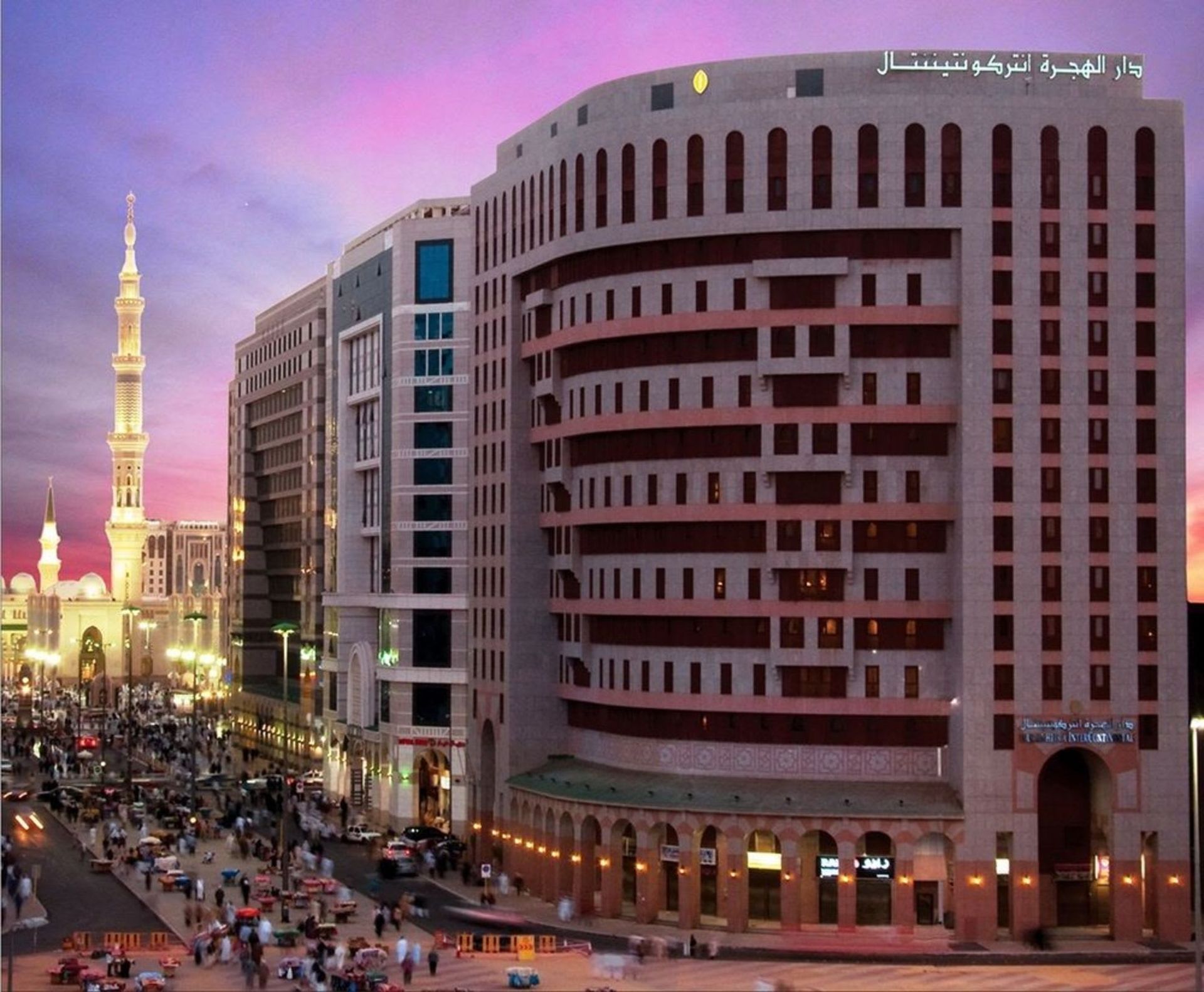 Large hotel buildings with arched windows, street with people, mosque minaret at sunset.