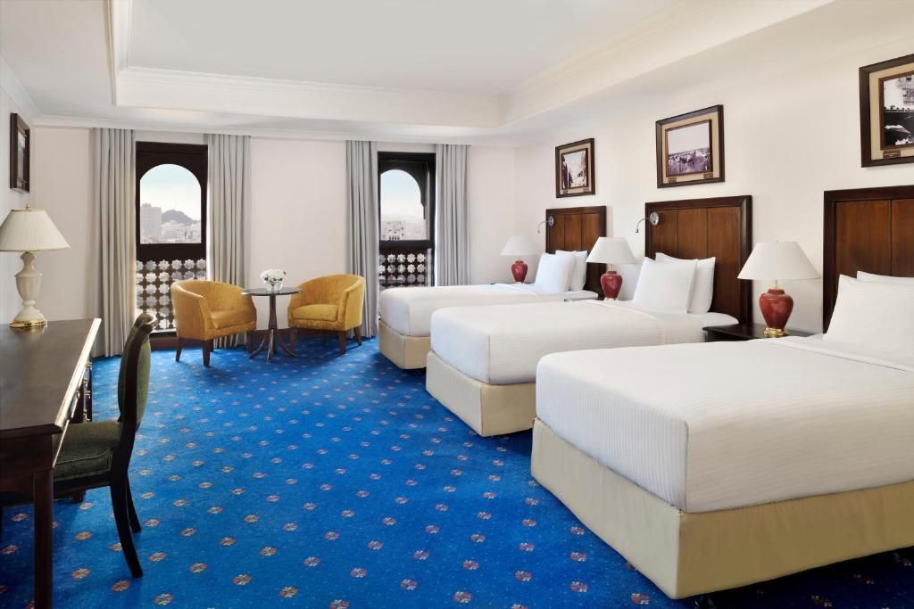 A hotel room with three beds, blue carpet, desk, and seating near arched windows with a city view.