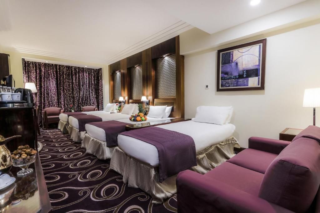 Hotel room with three beds, purple accents, and a sofa.
