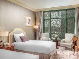 Hotel room with two twin beds, a decorative window, and two armchairs. Beige and light green colors.