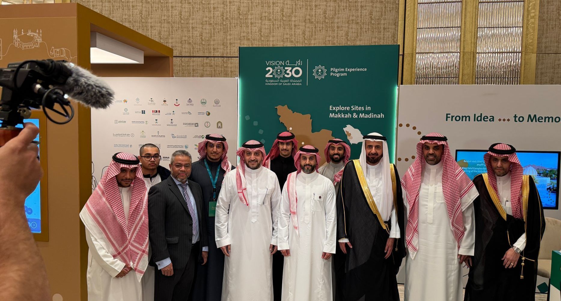 Group of men in traditional attire posing in front of a display with Arabic text and green background.