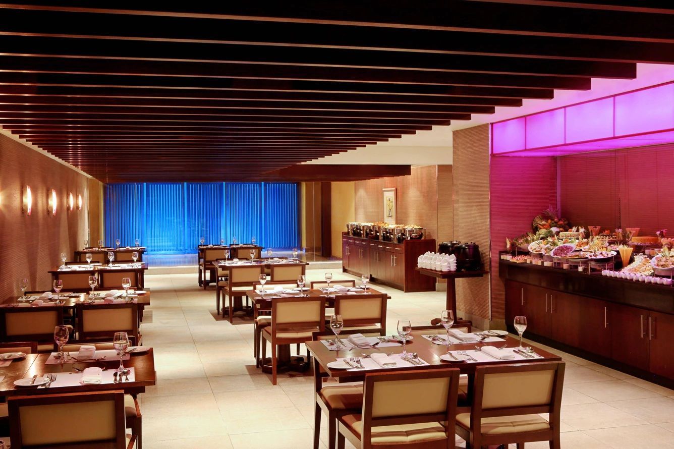 Restaurant interior: tables set for diners, buffet stations with food, wood and beige tones, blue curtain backdrop.