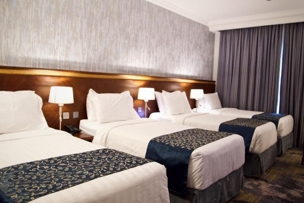 Four beds in a hotel room, white linens, gray and blue accents, neutral-toned walls, bedside lamps.