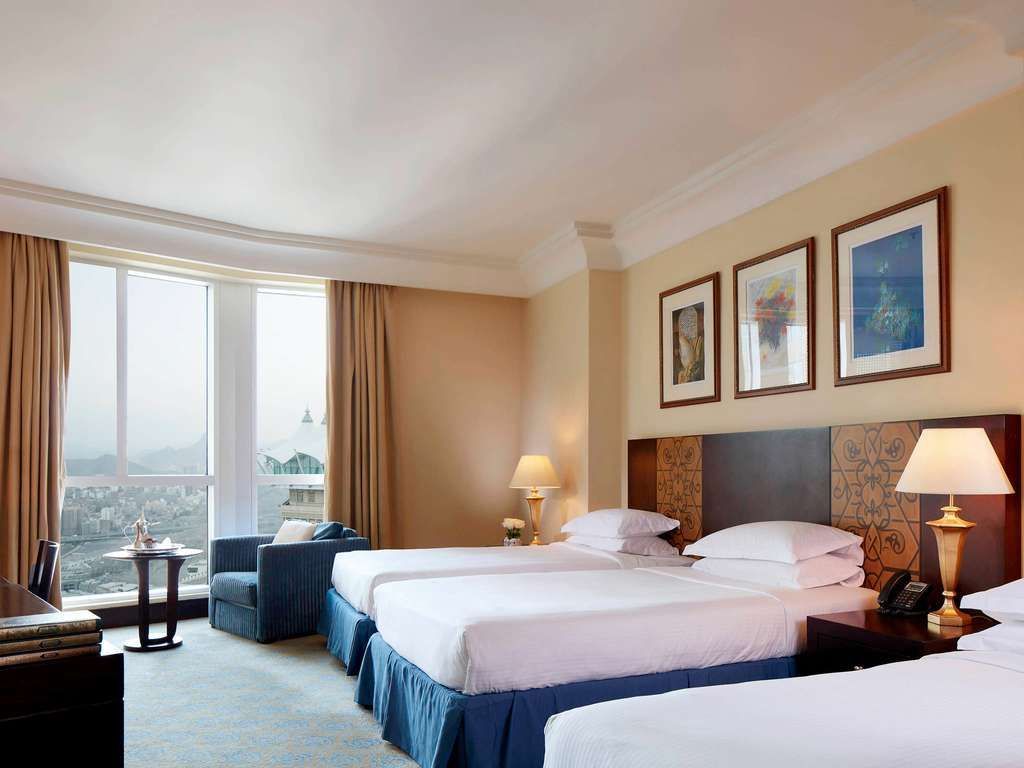 Hotel room with three beds, window with a view, armchair, lamps, and wall art.
