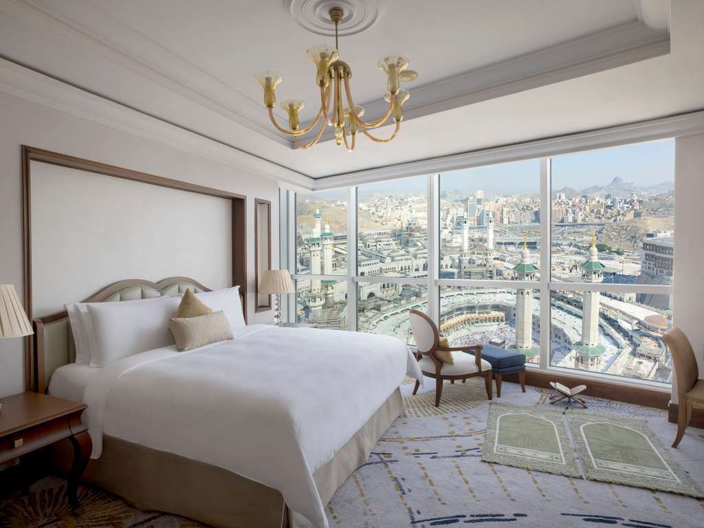 Hotel room with a bed, chair, prayer rug, and city view.