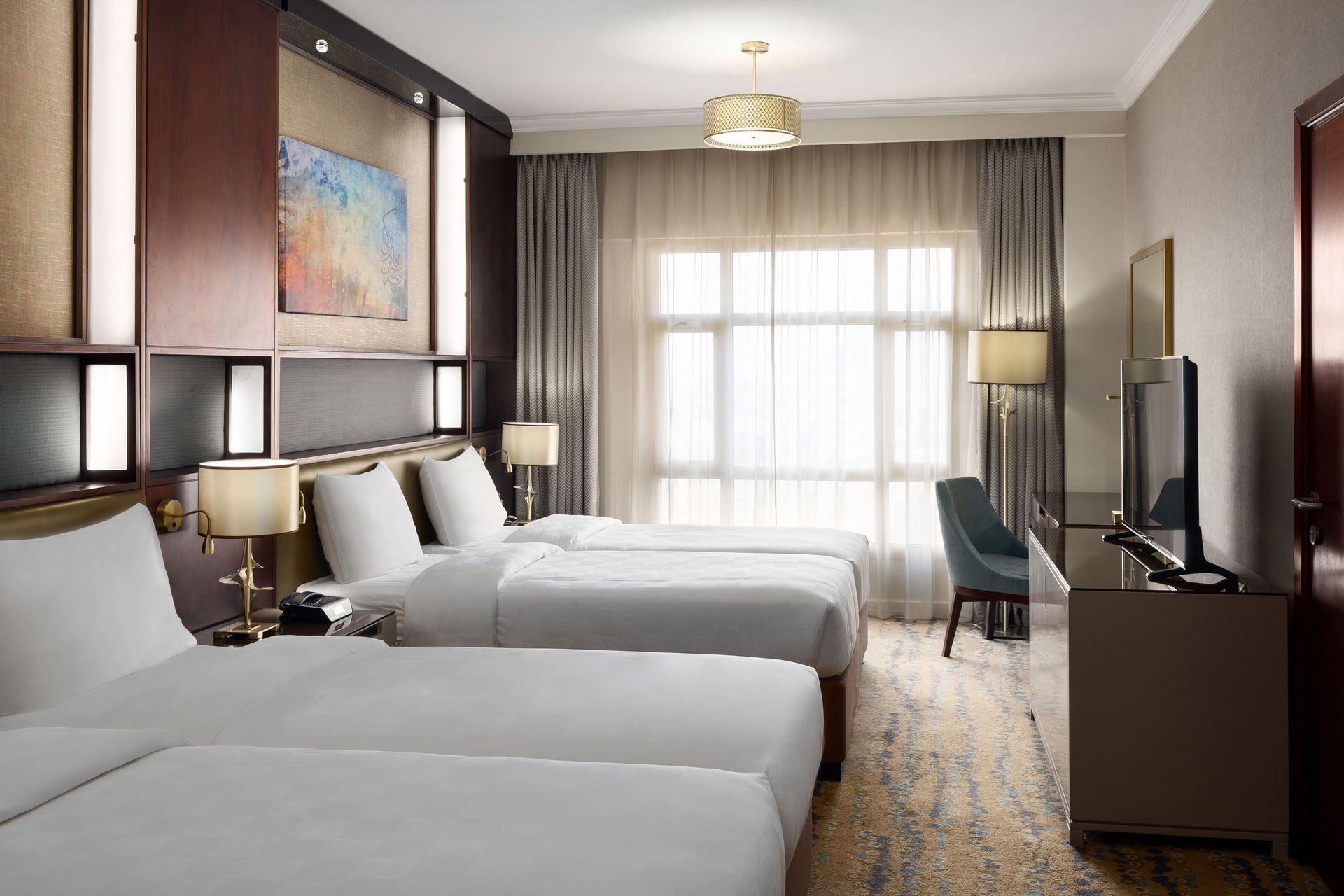 A hotel room with three beds, a window, and a desk. White linens, neutral tones.