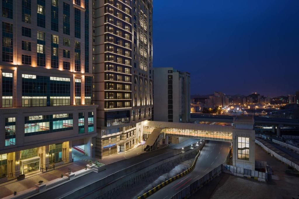 A modern hotel building lit up at night, with a pedestrian bridge and street.
