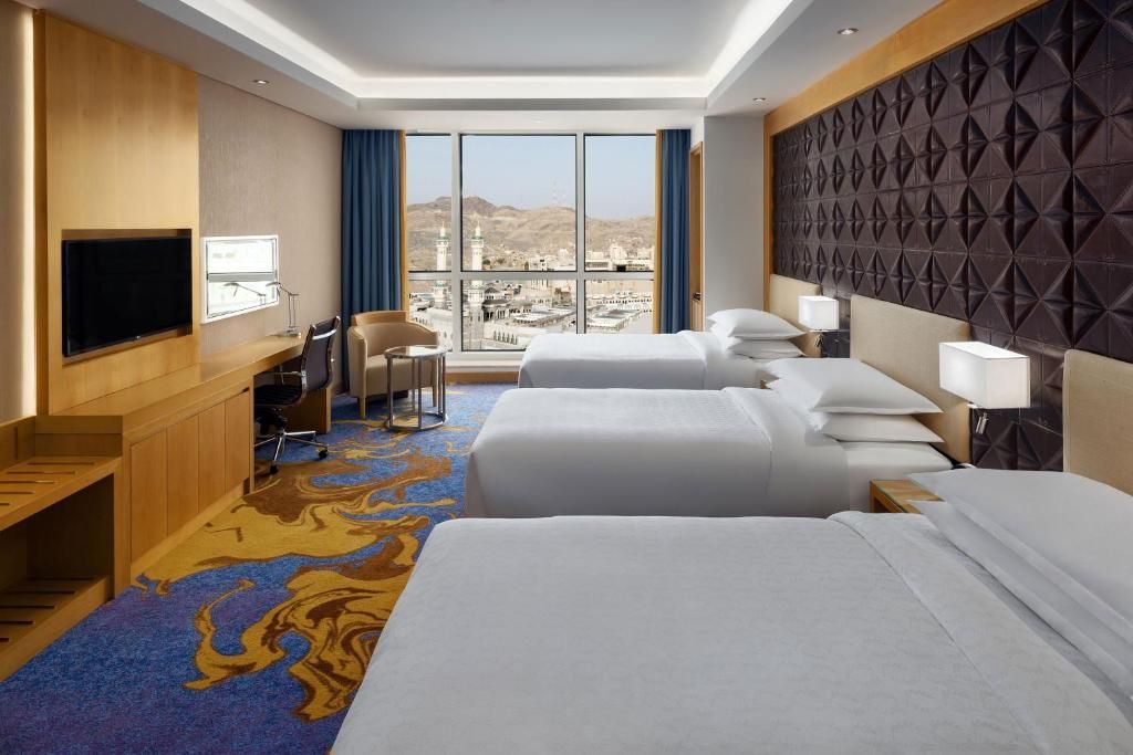 Hotel room with three beds, a desk, TV, and window overlooking a city.