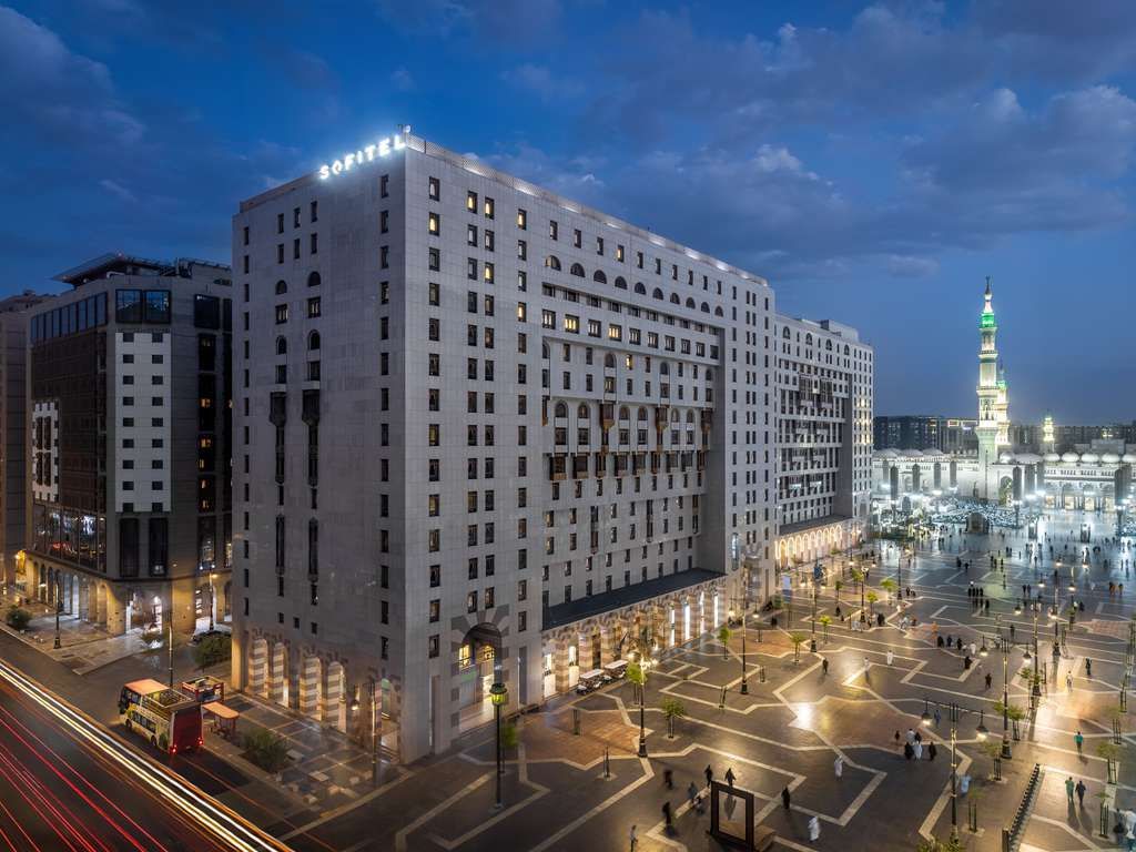 Exterior view of the white Dar Al-Taqwa Hotel, Medina, Saudi Arabia, at dusk, with people in the courtyard and mosque minaret in the distance.