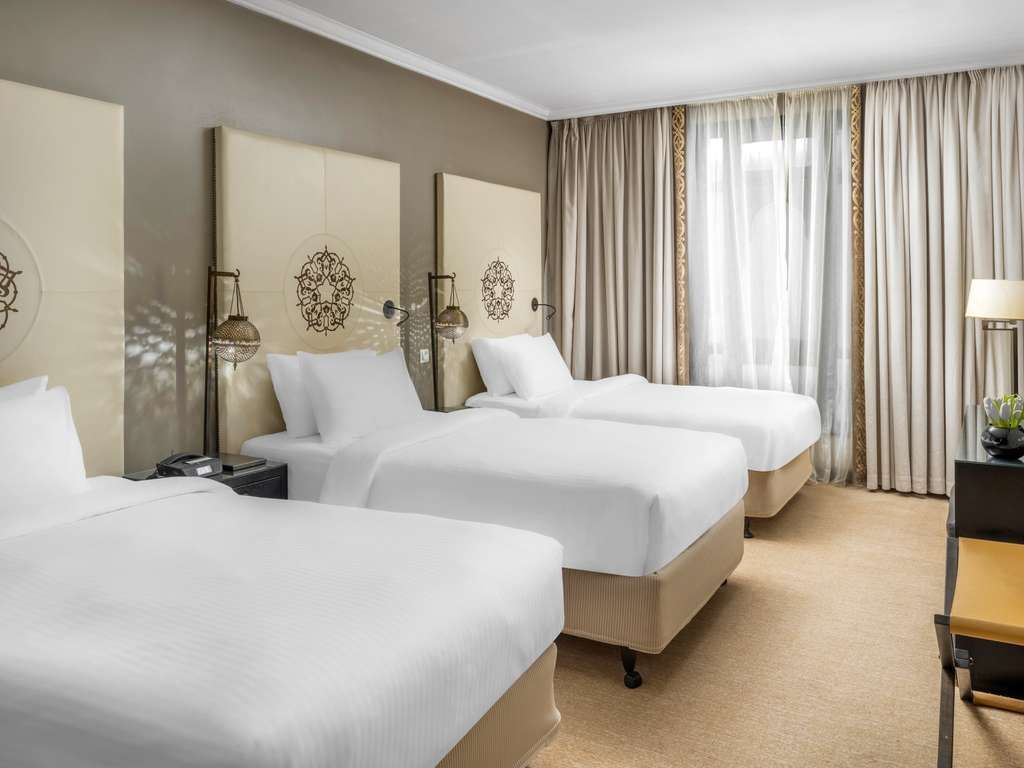 Hotel room with three beds, neutral color palette, ornate headboards, window with curtains.
