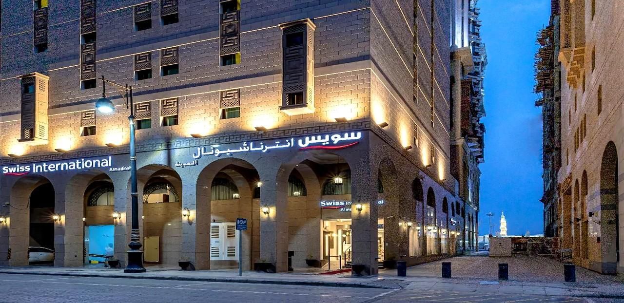 A building at night with Arabic text, streetlights, and a narrow alleyway leading to a tower.