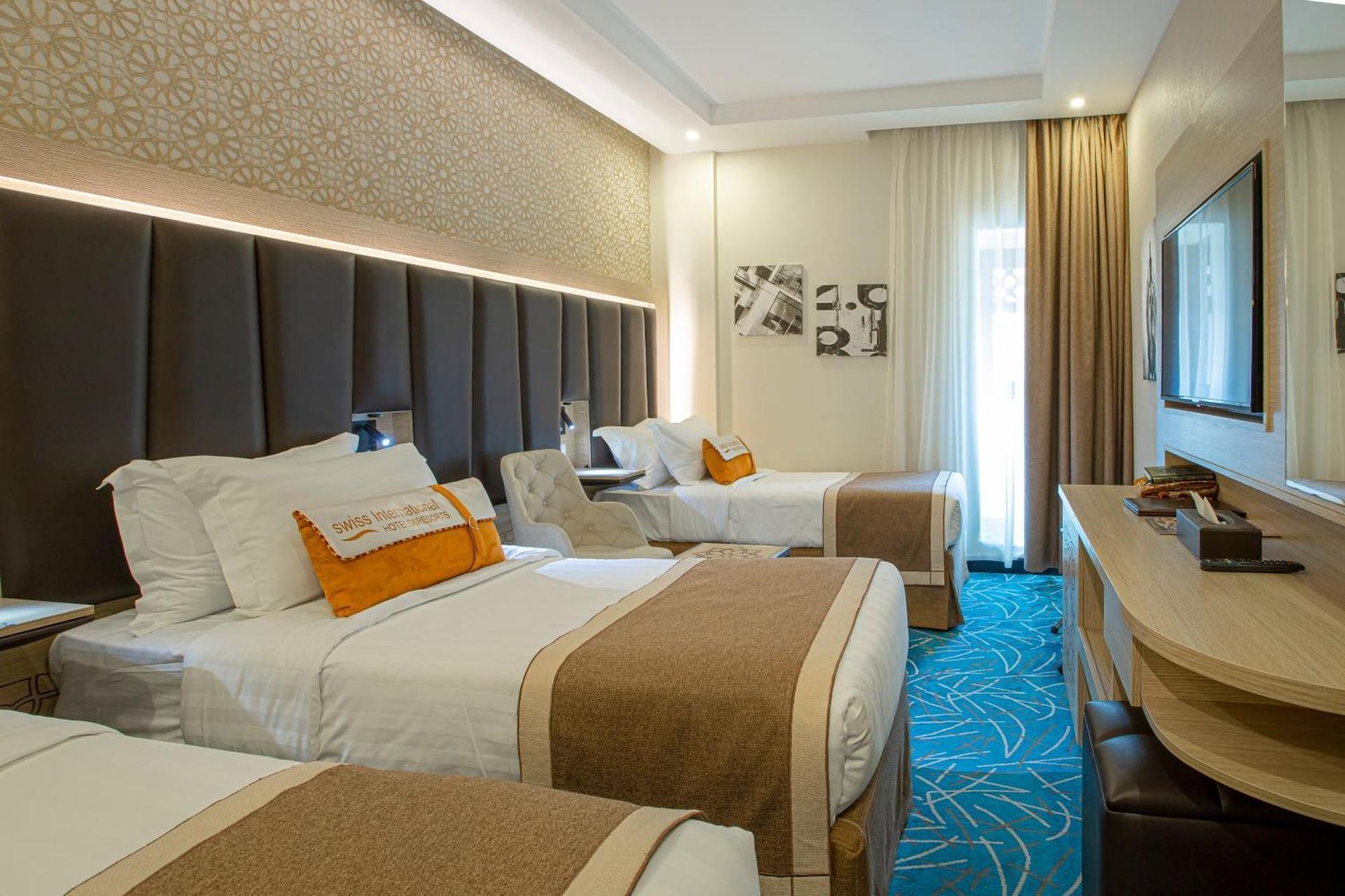Hotel room with three beds, brown and orange accents, TV, patterned blue carpet.