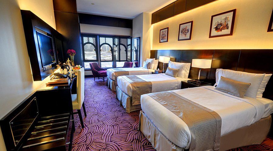 Hotel room with three beds, desk, and window with seating. Purple carpet, neutral bedding, and artwork.