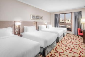 Hotel room with four beds, beige walls, patterned carpet, and a city view.