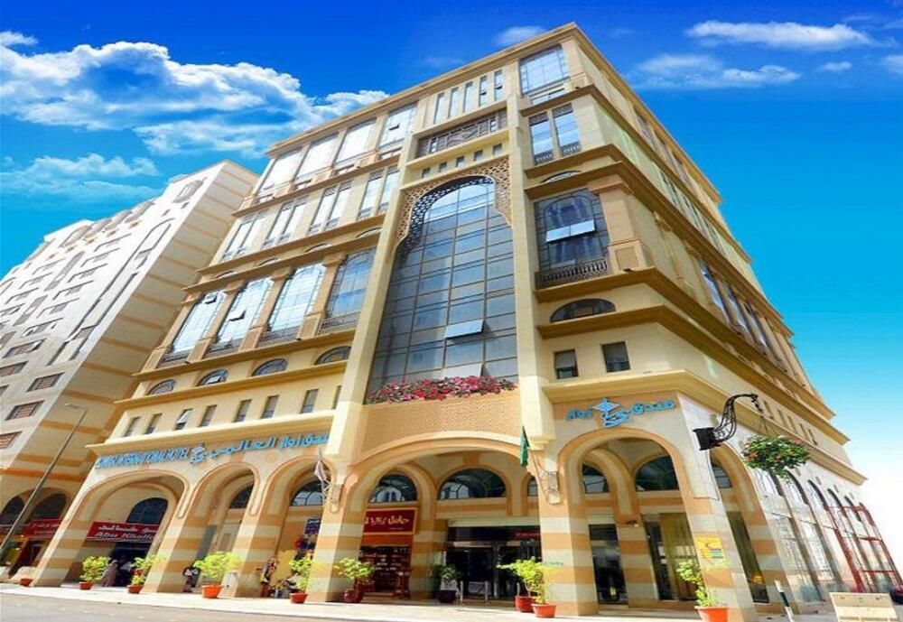 A beige and glass-fronted hotel building in a city, with arched entrance and sky with clouds.