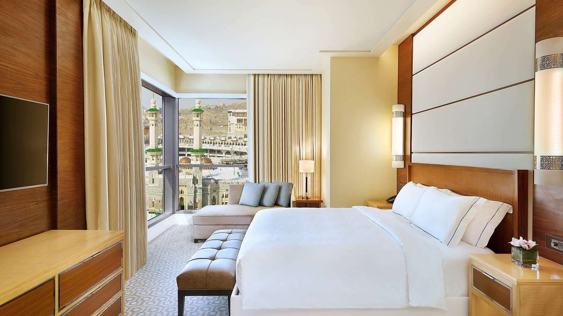 Hotel bedroom with a view of a city and a large bed. Beige and brown tones, natural light.