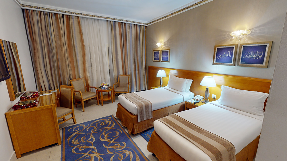 Twin beds in a hotel room, neutral colors with wood furniture, a blue rug, and artwork.