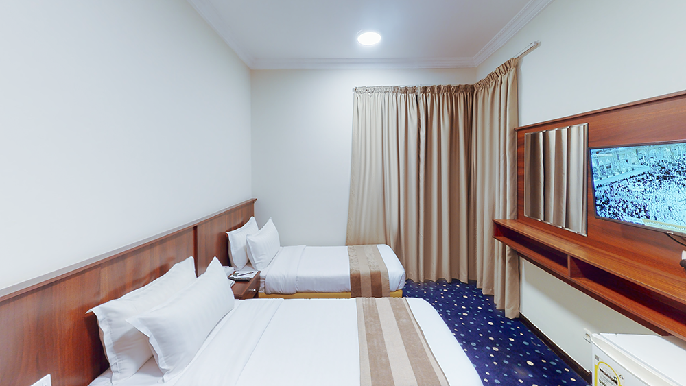 Hotel room with two beds, TV, and beige curtains against white walls and blue patterned carpet.