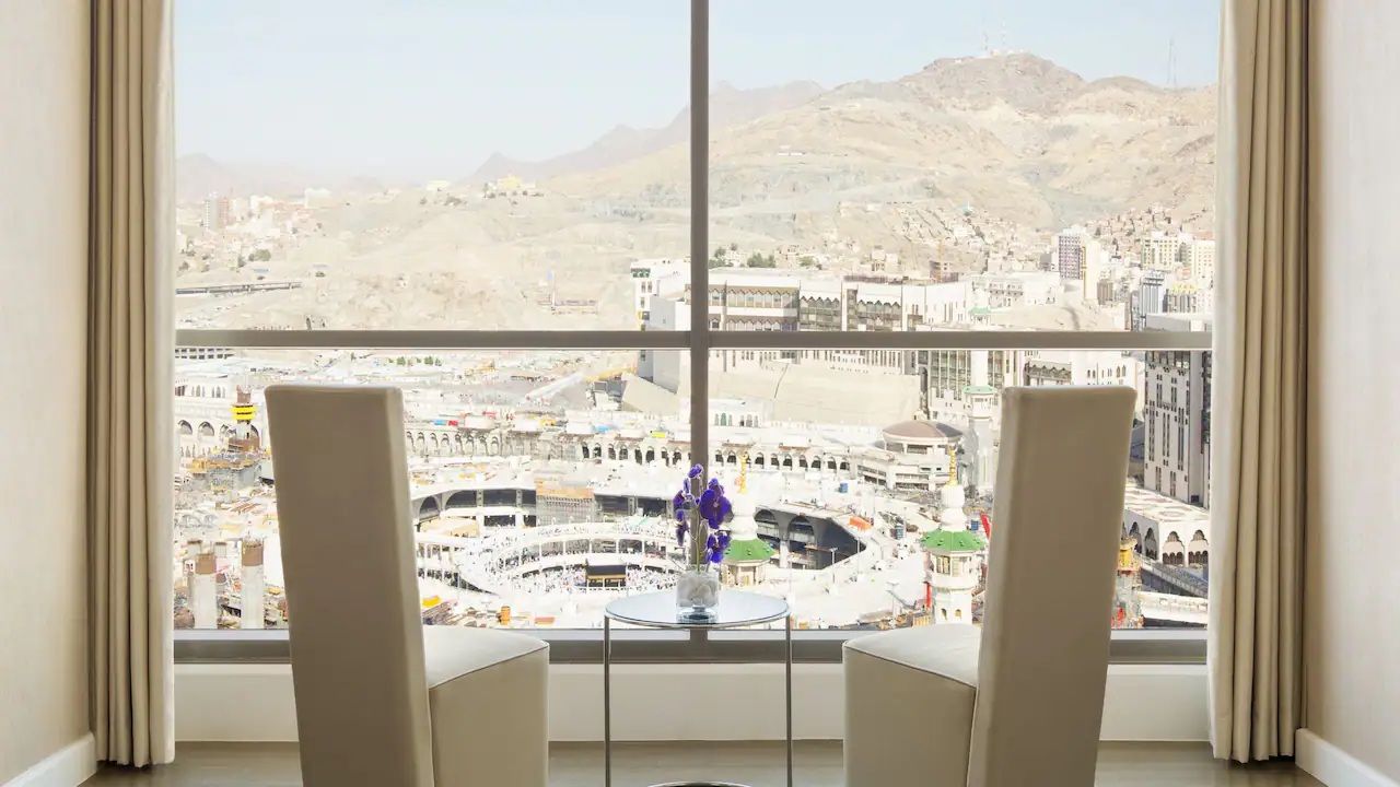 View of Mecca from a window with two chairs. White buildings and mountains in the background.