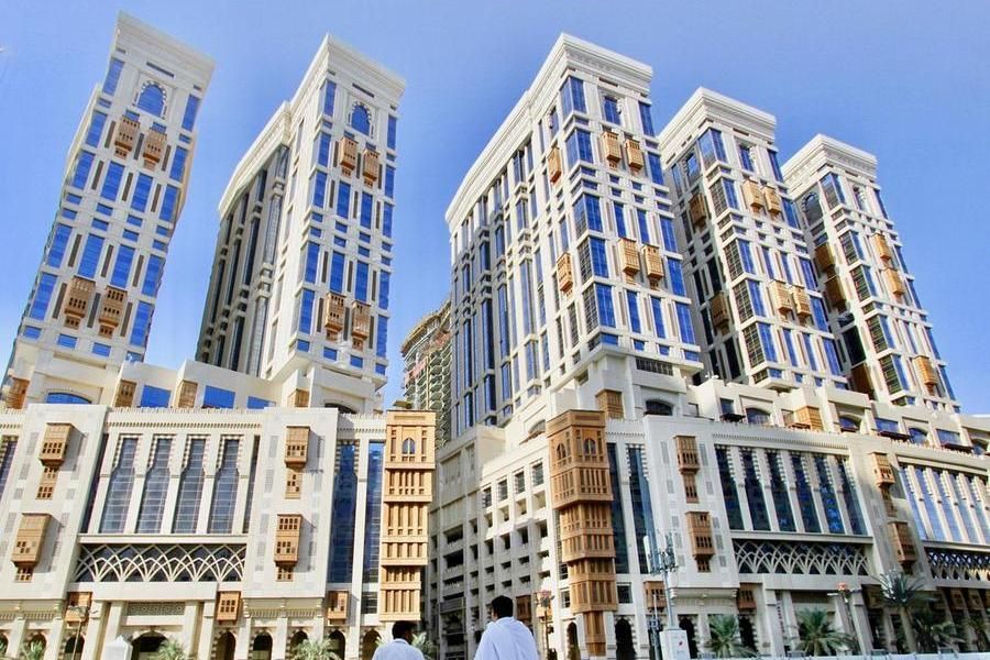 Tall white buildings with blue-tinted windows in Mecca, Saudi Arabia, with people walking in front.