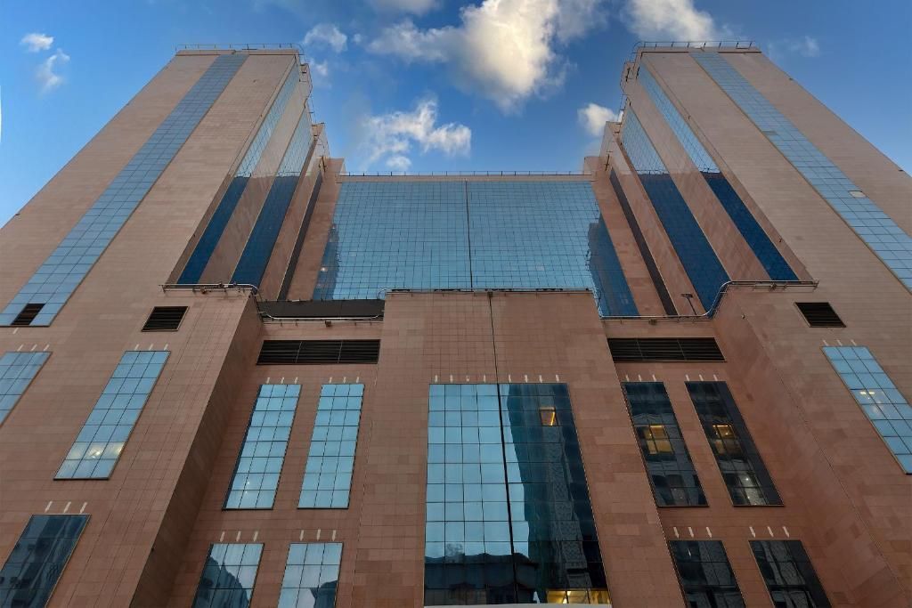 Tall brick and glass building, two towers connected by a lower central section, against a blue sky with clouds.