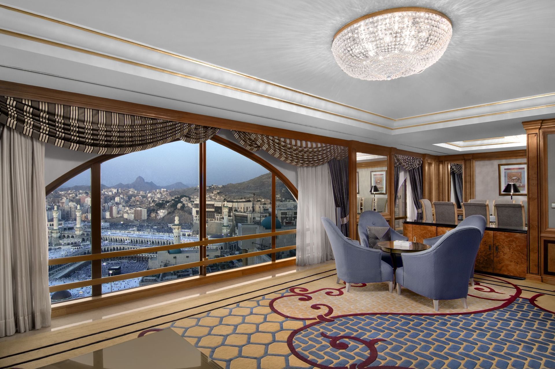 Elegant hotel room with city view, blue seating, ornate rug, and large window.