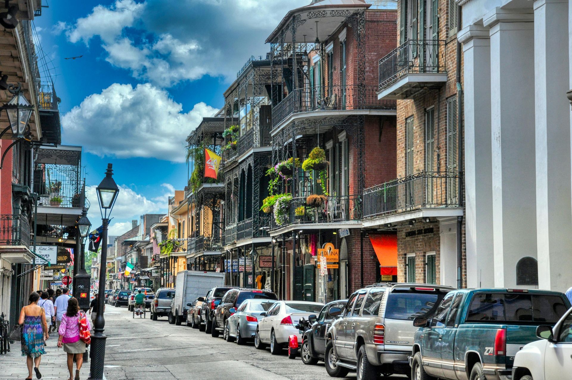 New Orleans street lined with parked cars and buildings with balconies; people walking.