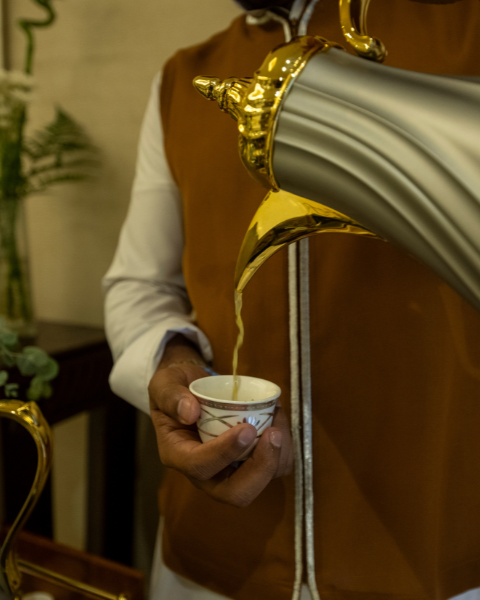 Person pouring coffee from a silver and gold pot into a small cup; brown vest, white shirt, and setting.
