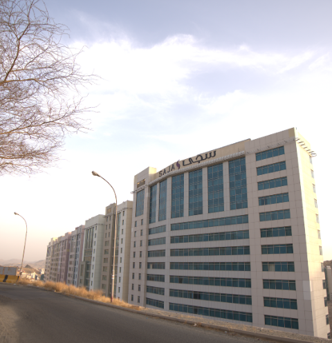 White high-rise building with blue-tinted windows and a sign in Arabic script, next to a road, under a cloudy sky.