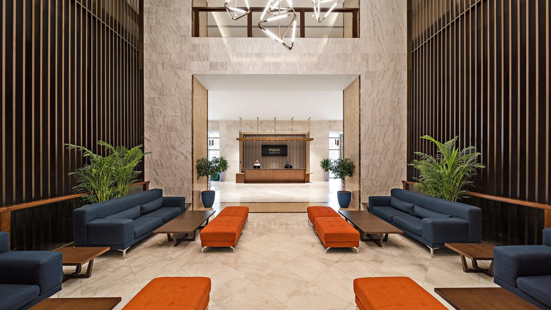 Hotel lobby: long shot. Blue couches and orange benches face each other on a marble floor. Reception desk in the back.