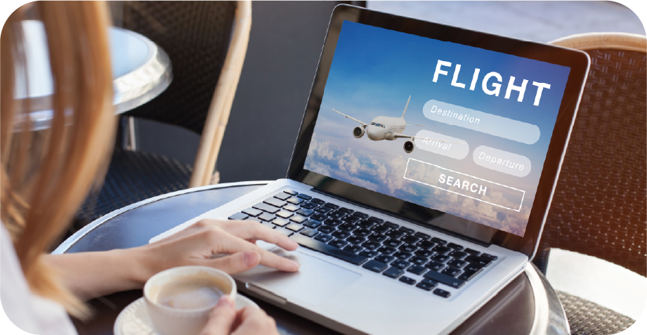 A woman is sitting at a table with a cup of coffee and using a laptop to search for a flight