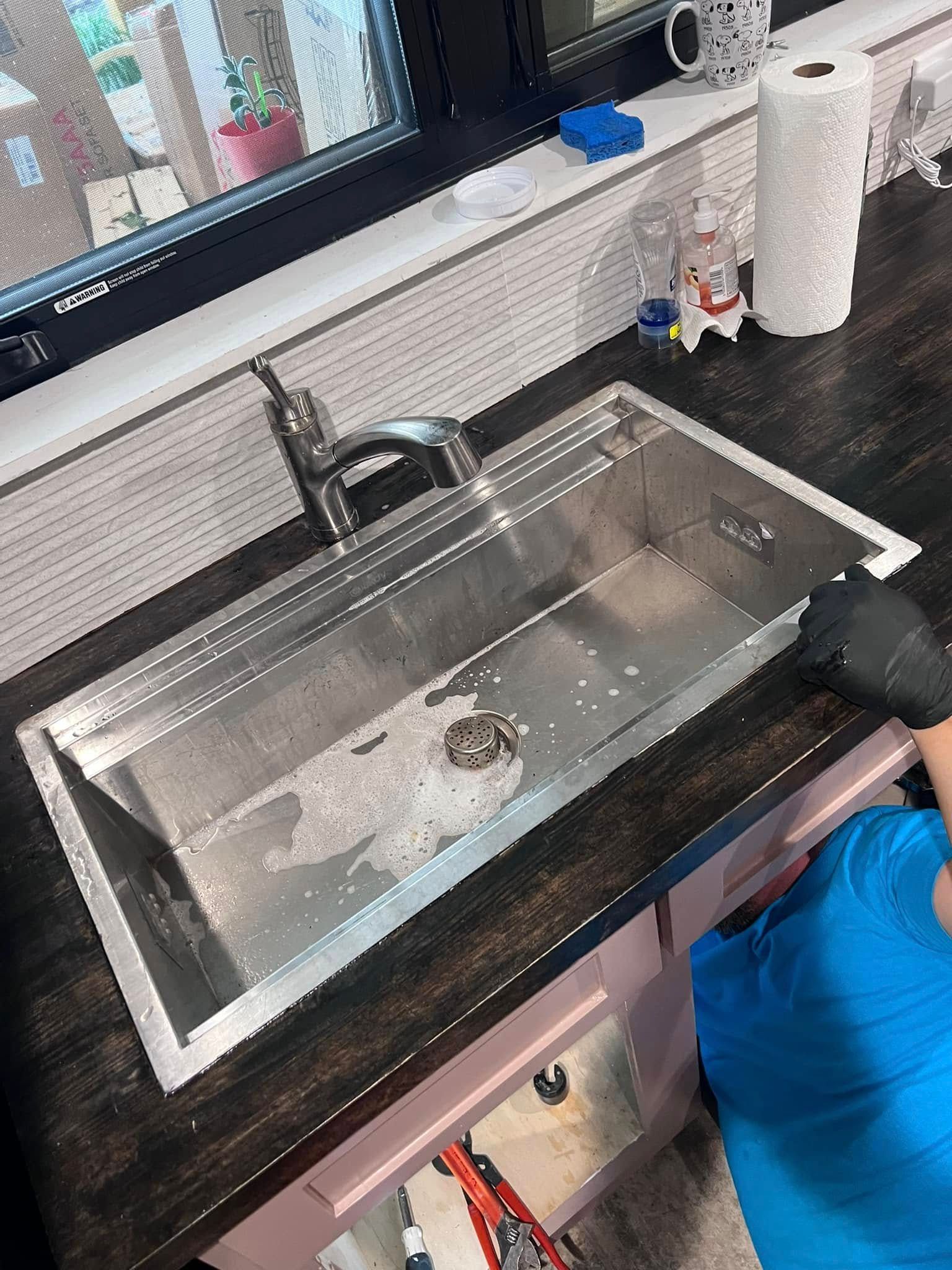 Stainless steel kitchen sink being cleaned, with a person’s gloved hand holding a sponge.
