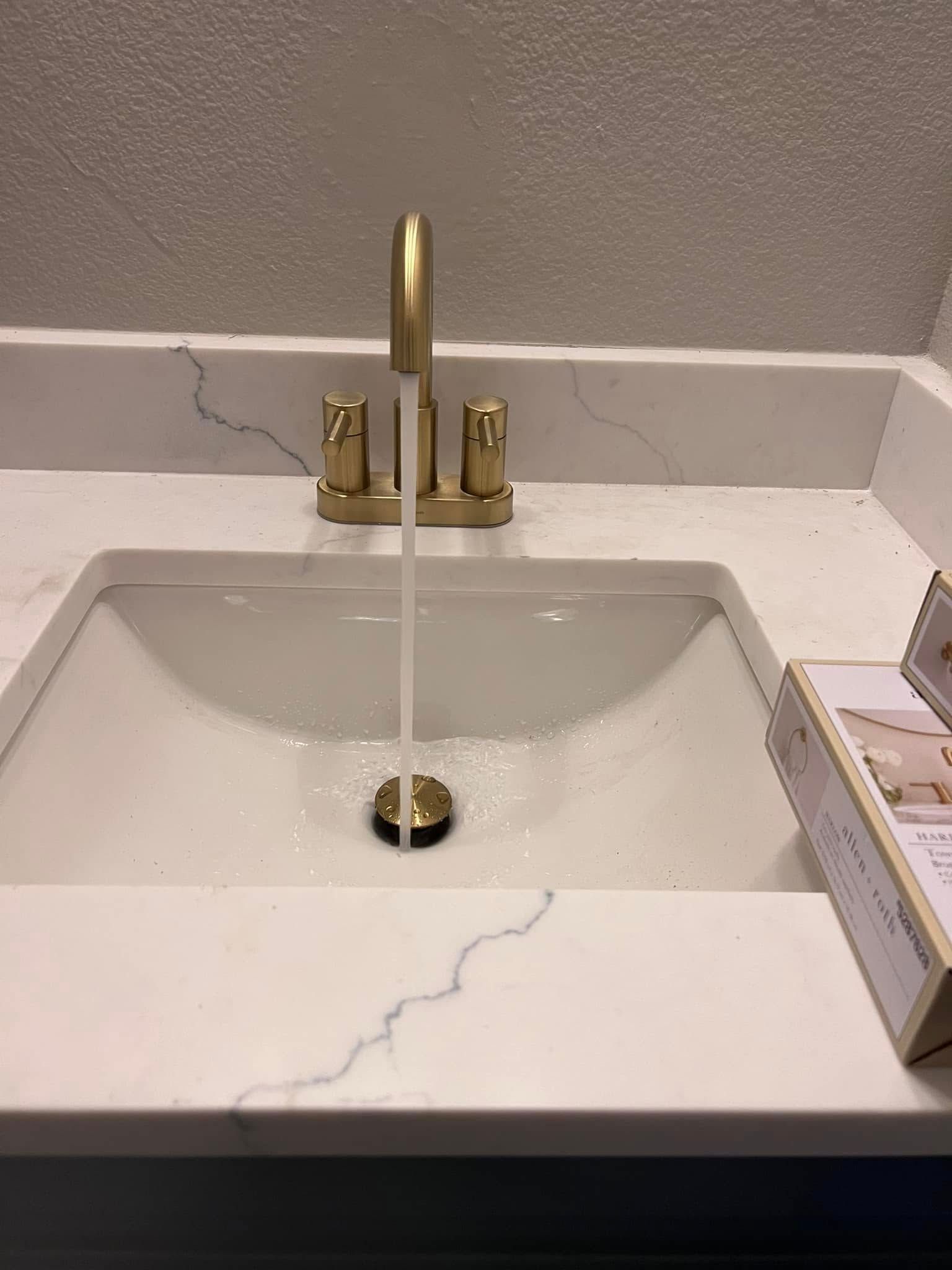 White marble bathroom sink with gold faucet running water and black drain.