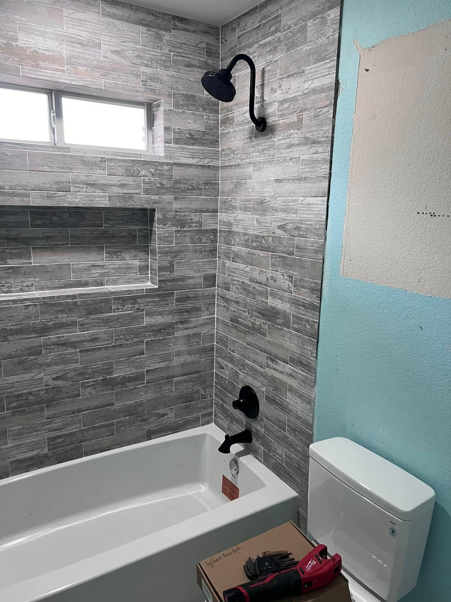 Small bathroom with gray tile, white tub, black showerhead, and a toilet beside the tub.