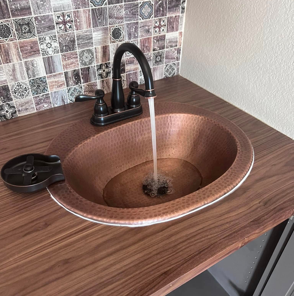 Copper sink with black faucet running water on a wood countertop with mosaic backsplash