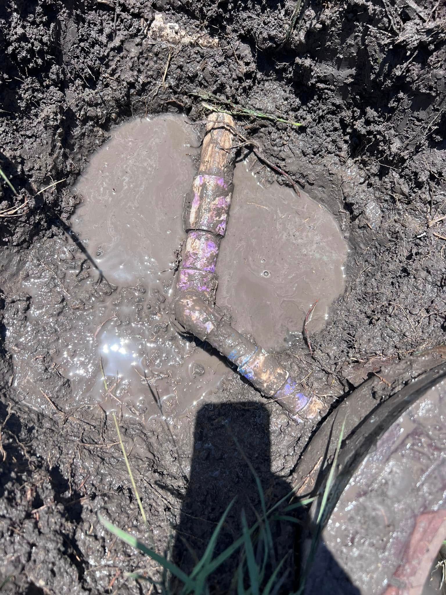 Gloved hand holds a rusty pipe over muddy ground with two wet concrete patches.