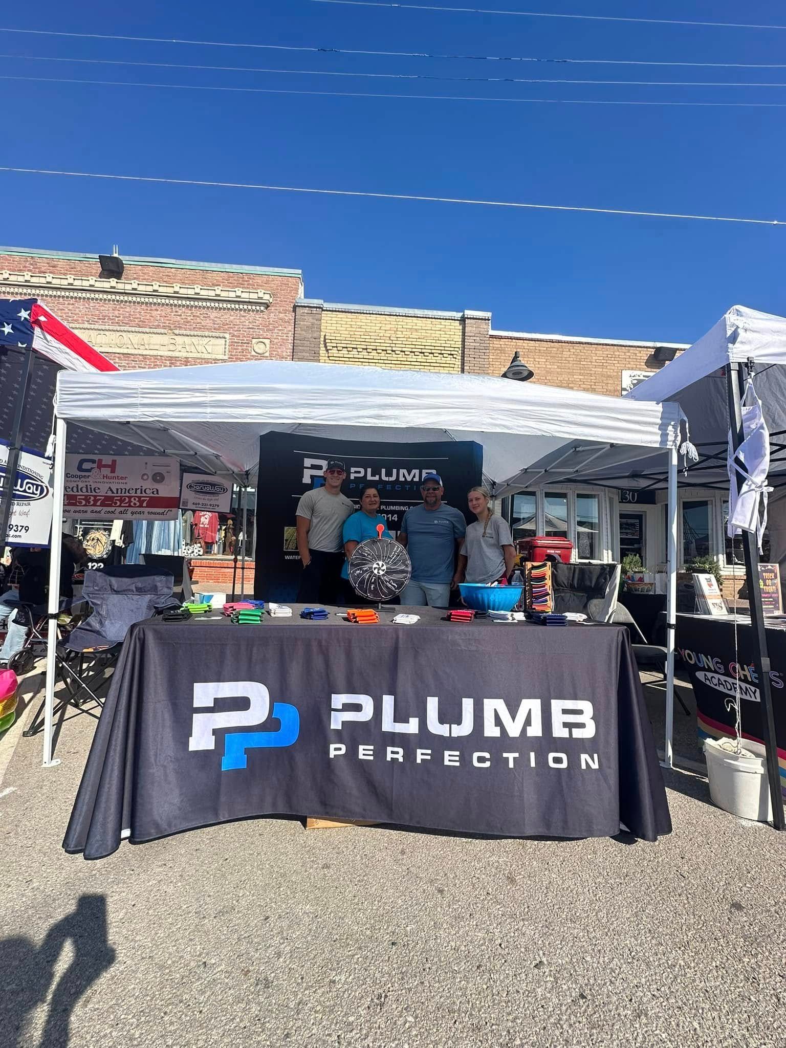 Plumb Perfection booth at an outdoor event with staff and branded table display.