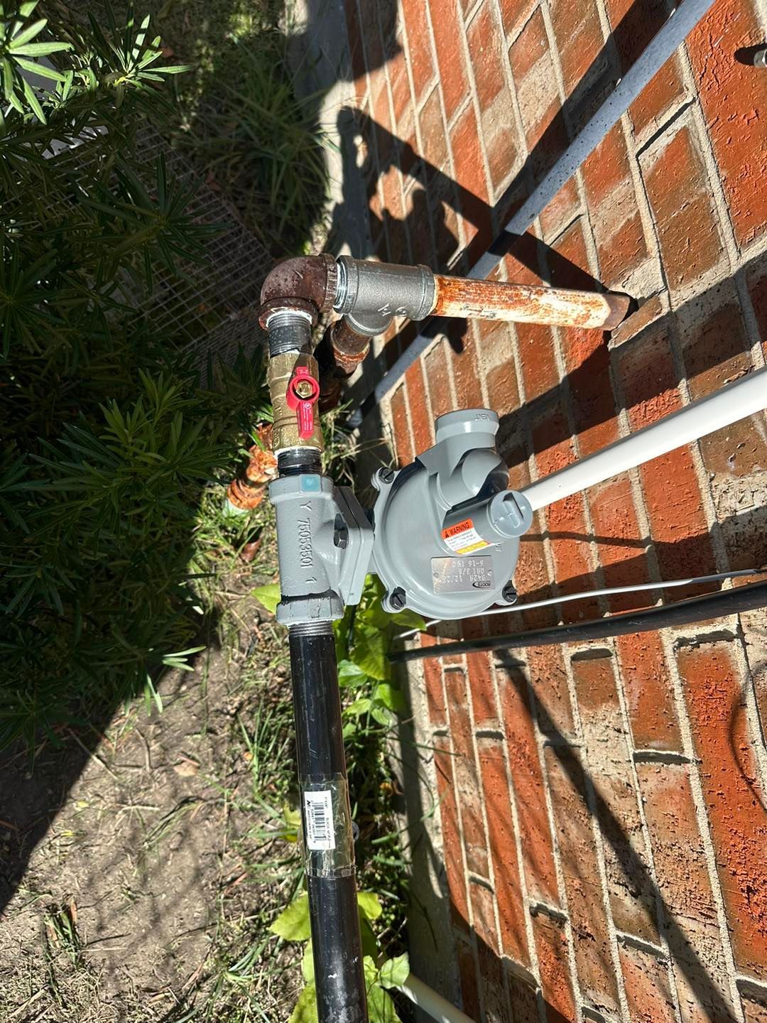 Outdoor gas meter and pipe assembly mounted beside a brick wall and grass.